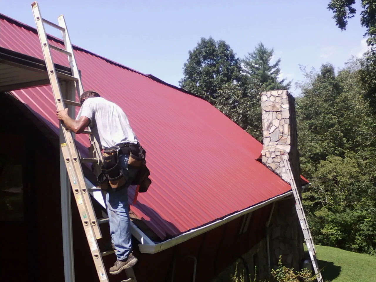 Roofer on a ladder beside a red metal roof and stone chimney — Soffit & Fascia Repair by Roofing Brothers in Gaston